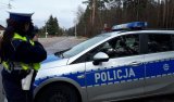 Zdjęcie w kolorze. Na pierwszym planie policjant z Wydziału Ruchu Drogowego KPP w Hajnówce mierzy prędkość pojazdów.  Na drugim planie zdjęcia widoczny jest radiowóz.