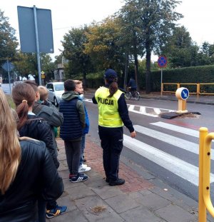 Dzieci wraz z Policjantką na edukacyjnym spacerze. Policjantka tłumaczy dzieciom jak prawidłowo przechodzić przez przejścia dla pieszych.