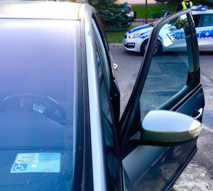 Samochód z otwartymi drzwiami, za szyba leży karta parkingowa dla osób niepełnosprawnych, w oddali stoi radiowóz policyjny